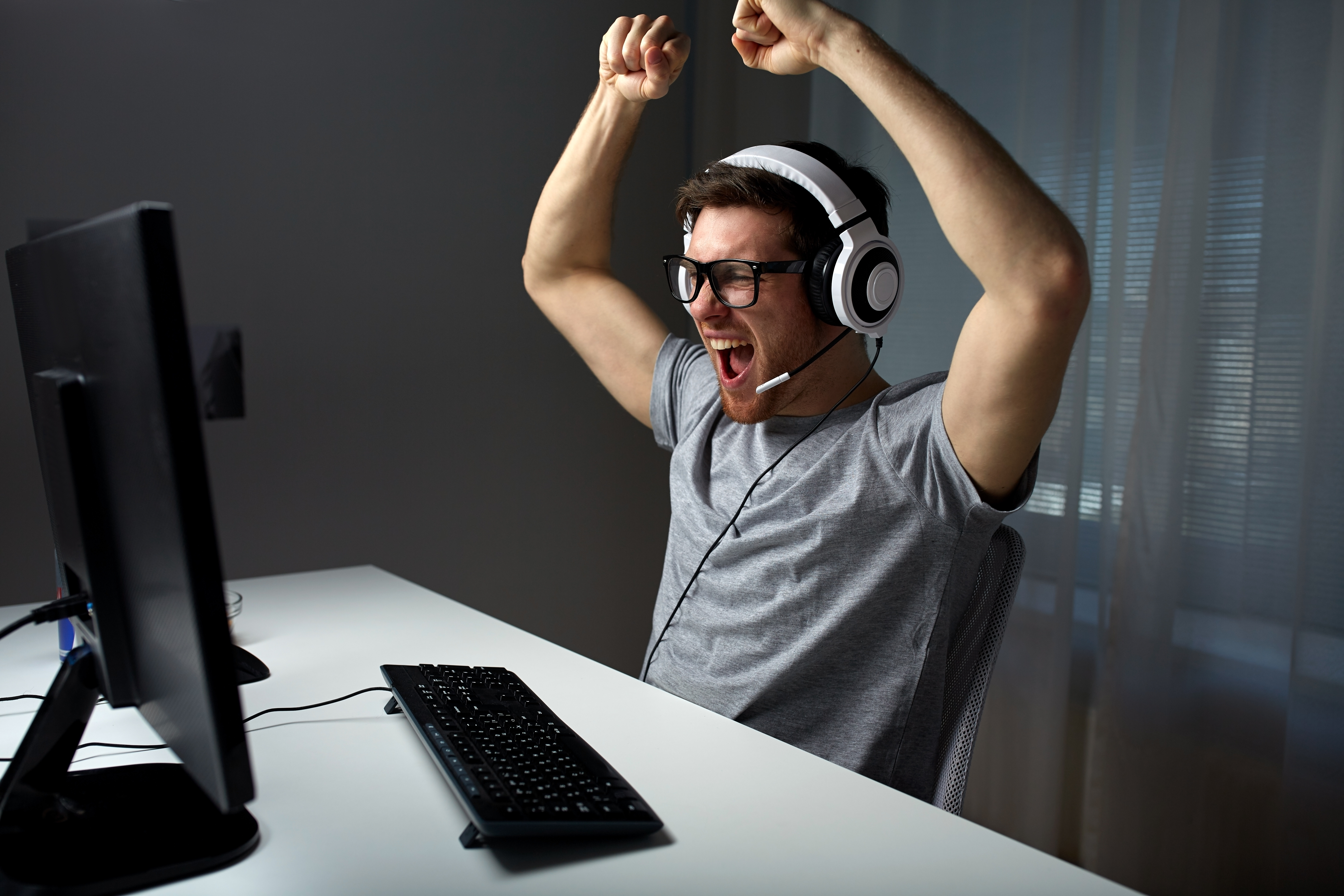 technology, gaming, entertainment, let's play and people concept - happy young man in eyeglasses with headset playing and winning computer game at home and streaming playthrough or walkthrough video